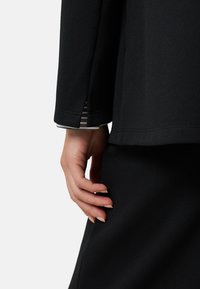 Czarny, teksturowany strój o luźnym kroju. Widoczna dłoń dotykająca ubrania, ukazująca pod rękawem paskowany akcent.