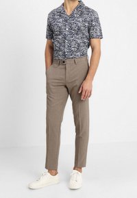 Man som bär en mönstrad kortärmad skjorta, slim-fit beige byxor med bälteshällor och vita sneakers, stående med en hand i fickan.