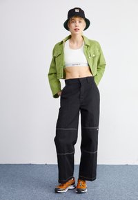 Veste courte en velours côtelé vert, débardeur blanc, pantalon noir à jambe large avec des coutures blanches, et baskets orange avec des accents noirs.