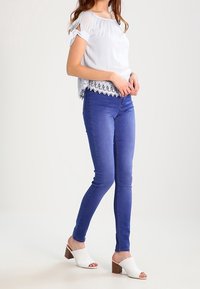 Blusa bianca con orlo in pizzo, abbinata a jeans skinny blu e calzature aperte bianche con tacco a blocco.