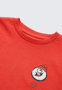 Rotes Baumwoll-T-Shirt mit einem runden, gestickten weißen Charakter, der eine Mütze trägt, mit dem Text „STAY WARM, STAY COOL“ darunter.