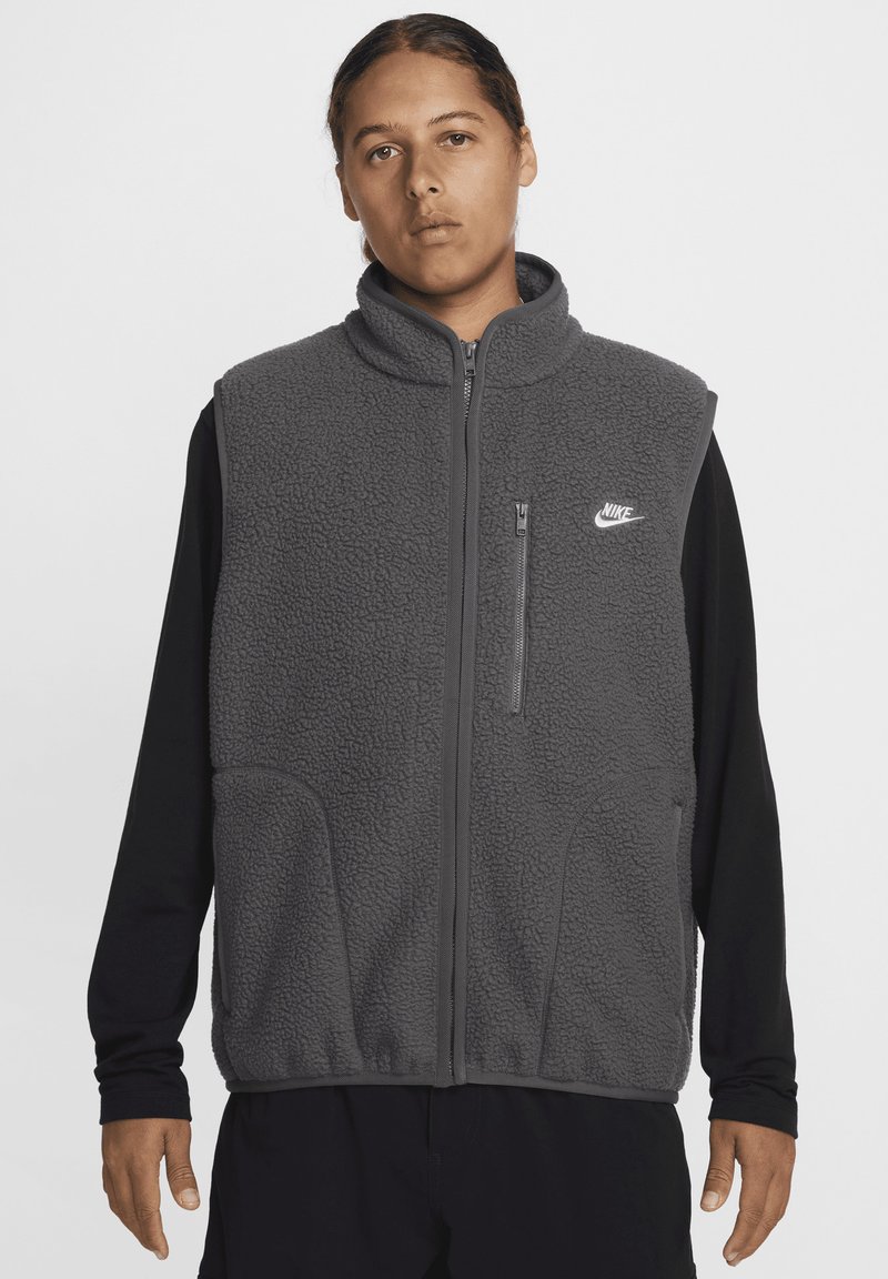 Pelēks vilnas bezdelīgains ar augstu apkakli, rāvējslēdzēja priekšpusi un sānu kabatām. Iezīmēts ar Nike logo kreisajā krūtīs un teksturētu audumu.