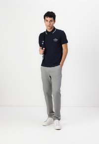 Marineblå polo med hvite krageakenter, grå slim-fit bukser og hvite sneakers. Modellen holder en marineblå veske. Enkel design, minimale mønstre.