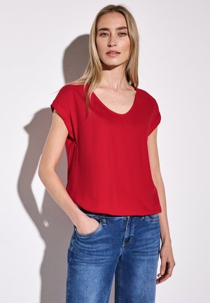 Maglietta rossa a maniche corte con scollatura a V, realizzata in tessuto morbido, abbinata a jeans blu, che evidenziano un fit rilassato e uno stile casual.