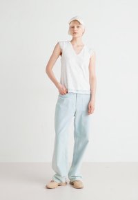 Hvit blonde topp med heftesærmer, V-hals, kombinert med lyseblå vidjeans. Modellen har på seg beige slip-on sko og en hvit caps.