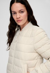 Doudoune beige clair avec fermeture éclair à l'avant, col côtelé et poches latérales. Présente des coutures horizontales et une finition lisse et mate.