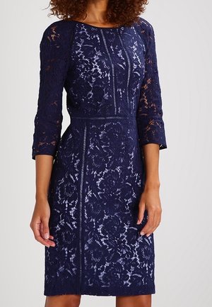 Robe en dentelle bleu marine avec manches trois-quarts, taille cintrée, motif floral et ourlet arrivant au genou portée par une personne aux cheveux bouclés.