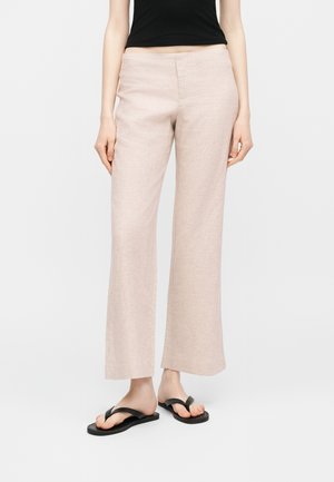 Άτομο που φοράει μπεζ παντελόνι με φαρδιά μπατζάκια, μαύρο αμάνικο τοπ και μαύρα σανδάλια, στέκεται μπροστά σε απλό ανοιχτόφωτο φόντο.