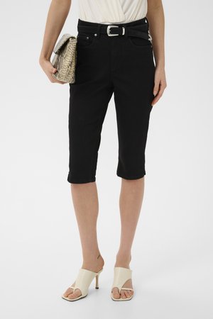 Frau trägt schwarze knieengebundene Shorts mit einem schwarzen Gürtel, hält eine Clutch aus Schlangenhaut und trägt weiße Sandaletten mit hohen Absätzen.