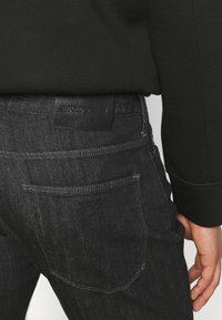 Mörka jeans i denim med en svart läderetikett vid midjan, med subtila sömnadsdetaljer och en liten logga präglad på bakfickan.