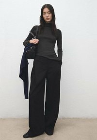 Black ribbed turtleneck paired with loose-fitting black wide-leg trousers. A navy jacket draped over one arm; a small bag in hand.