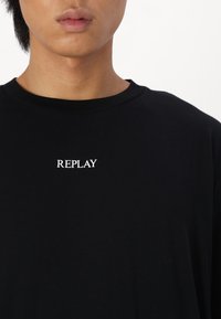 Černé bavlněné tričko s kulatým výstřihem a bílým logem "REPLAY" na hrudi. Měkká textura, volný střih, minimalistický design.