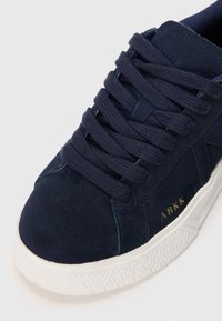 Tenis navy din suede cu limbă texturată, șireturi plate navy, talpă din cauciuc albă și logo-ul „ARKK” embosat de culoare aurie pe laterale.
