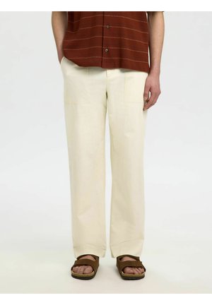 Personne portant un pantalon crème à jambes droites, des sandales marron à bout ouvert, et une chemise à manches courtes couleur rouille avec fines rayures.