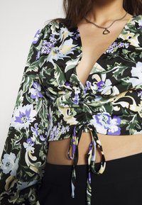 Top corto floreale con scollo a V profondo, caratterizzato da motivi verdi, viola e gialli su uno sfondo nero; arricciature frontali e dettaglio con fiocco all'orlo.