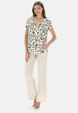 Donna con camicetta corta a maniche corte con scollo a V floreale color crema, pantaloni beige a gamba larga e scarpe bianche con tacco alto, in piedi con una mano in tasca.