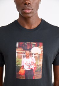 Schwarzes Baumwoll-T-Shirt mit einem Frontdruck eines Basketballspielers, der einen Ball und Schuhe hält, mit roten und weißen Akzenten im Design.