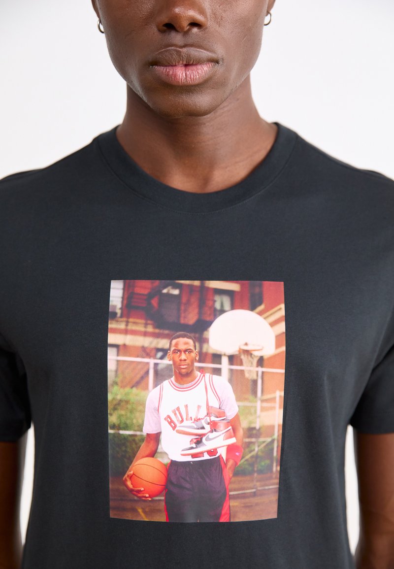 Czarny bawełniany T-shirt z nadrukiem z przodu przedstawiającym koszykarza trzymającego piłkę i buty, z czerwonymi i białymi akcentami w projekcie.