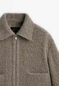 Ruskea villatakkia karkealla pinnalla, vetoketjulla edessä ja kauluksella. Siinä on kaksi etutaskua ja hienovaraiset ompelu- ja tikkaukset.