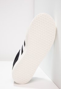 Zapatilla negra con una suela de goma blanca texturizada. El detalle lateral presenta tres franjas negras paralelas sobre un material superior oscuro.