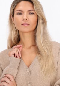 Beige Strickjacke mit V-Ausschnitt und zwei vorderen Knöpfen. Sanfte Textur mit gerippten Bündchen. Die Haare sind lang und blond, glatt gestylt.