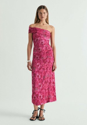 Mujer con un vestido midi sin mangas, rosa con estampado, con detalle de hombros descubiertos y sandalias negras con cordones, de pie frente a un fondo blanco liso.