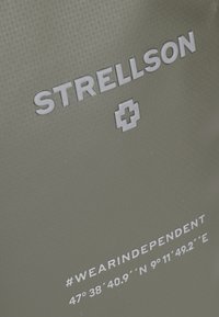 Borsa verde oliva con logo "STRELLSON" in rilievo e testo bianco. Presenta coordinate geografiche e hashtag "#WEARINDEPENDENT". Superficie testurizzata.