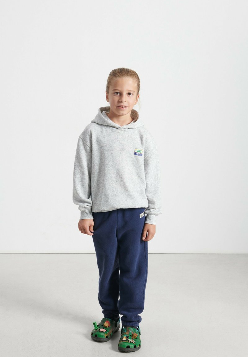 Felpa grigia con cappuccio, tasca frontale e patch con logo, abbinata a pantaloni della tuta blu navy e sandali verdi con design di animali.