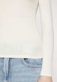 Maglia a maniche lunghe a coste color crema con un design aderente, caratterizzata da un logo discreto sull'orlo, abbinata a jeans in denim blu chiaro.