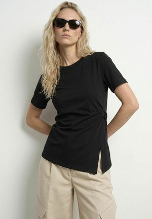Blonde vrouw die een zwart t-shirt met korte mouwen en een zijsplit draagt, en beige cargobroek, staand tegen een witte achtergrond met een zonnebril.