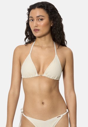 Mujer con cabello largo y rizado, vistiendo un bikini texturizado color blanco hueso, con top y parte inferior, sobre un fondo claro y liso.