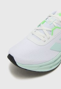 Zapatilla deportiva blanca con una parte superior de malla texturizada, detalles en verde, cordones planos y una suela gruesa en verde menta. Detalle de la suela exterior en negro visible.