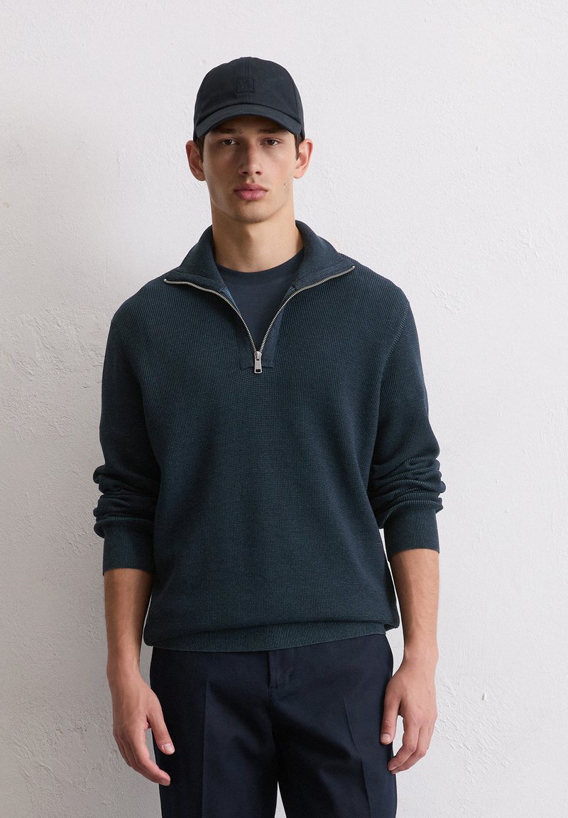 Ein dunkelgrüner Strickpullover im Einzelhandel, mit einem Reißverschlusskragen, langen Ärmeln und einem gerippten Saum. Getragen über einem dunklen T-Shirt, kombiniert mit dunklen Hosen.