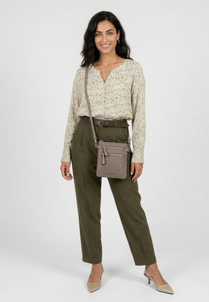 Woman with dark hair wears speckled beige blouse, olive high-waist trousers, beige heels, and crossbody taupe purse, smiling against white background.