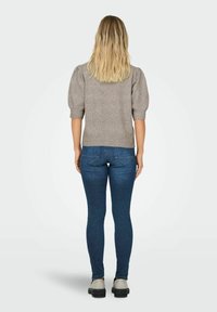 Vrouw met lang blond haar, gekleed in een grijze trui met korte mouwen, blauwe skinny jeans en witte schoenen, staat met haar rug naar de kijker.