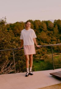 Jeune femme en chemise blanche et jupe rose, souriante, debout sur la rambarde d'une terrasse avec vue sur un paysage verdoyant et boisé.
