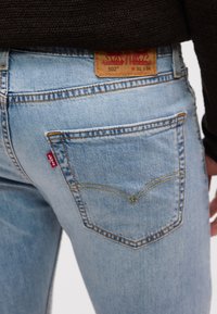 Πίσω τσέπη ανοιχτό μπλε τζιν με κόκκινη ετικέτα Levi's και καφέ δερμάτινο σήμα επωνυμίας στη μέση, φορεμένα με σκούρο πουλόβερ.