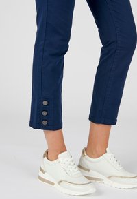 Marineblauwe cropped broeken met drie metalen knopen aan de zoom, gecombineerd met witte sneakers met gouden accenten en een gestructureerd oppervlak.