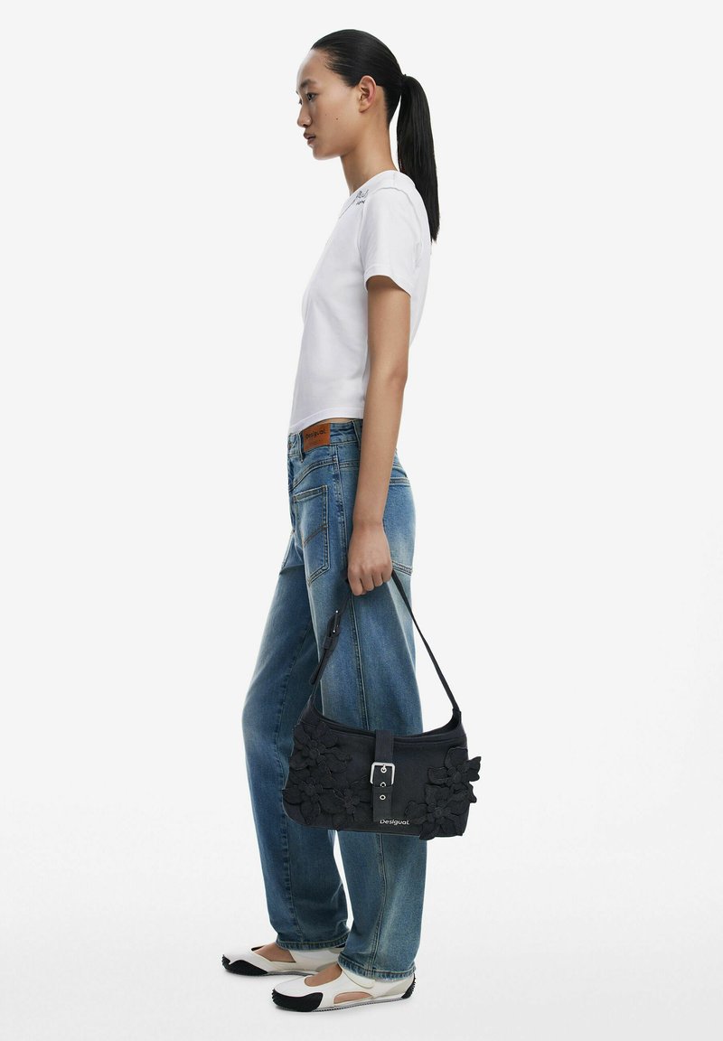 Black handbag with floral fabric embellishments, featuring a silver buckle and a shoulder strap. Model wears a white shirt and blue jeans.