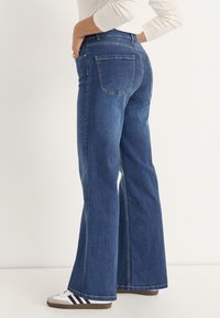 High-waisted, flared blauwe spijkerbroeken hebben een vijf-pocket ontwerp, contraststiksels en een soepele textuur, gecombineerd met witte sneakers.