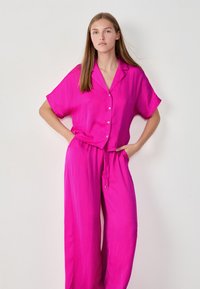 El conjunto de dos piezas fucsia presenta una blusa de botones de manga corta y pantalones de corte holgado con cintura elástica y cordón. Textura suave.
