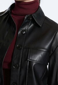 Veste en cuir noire avec col, boutons sur le devant et une poche poitrine. Portée sur un col roulé en maille bordeaux. Texture lisse.