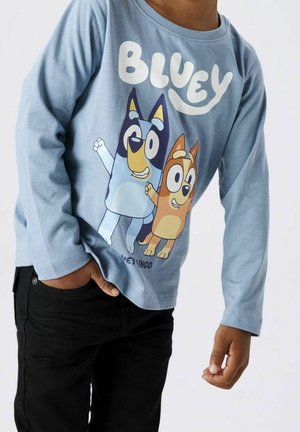 Barn iført blå langærmet trøje med tegnefilm Bluey og Bingo hunde og sorte bukser, den ene hånd i lommen og den anden afslappet.