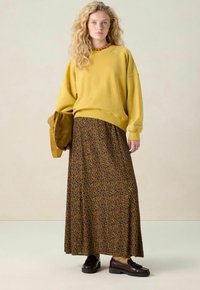 Femme aux cheveux blonds bouclés portant un pull jaune moutarde, une jupe longue à imprimé léopard, des mocassins marron et tenant un chapeau en suède marron.