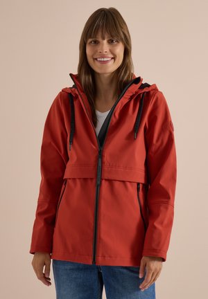 Femme souriante portant une veste à capuche rouge avec une fermeture éclair noire et un jean bleu sur un fond beige uni.