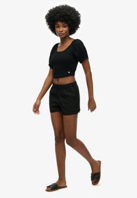 Zwart crop top met korte pofmouwen, gecombineerd met zwarte shorts met elastische tailleband. Model draagt slidesandalen. Eenvoudige achtergrond.