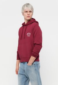 Hoodie bordeaux avec poche avant, capuche à cordon, et texte imprimé blanc sur la poitrine. Associé à un jean déchiré bleu clair.