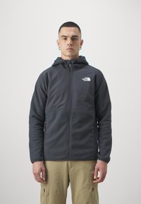 Kurtka typu softshell z kapturem w ciemnoszarym kolorze, z zamkiem z przodu, kontrastującą dolną częścią z polaru oraz logo na lewej piersi.