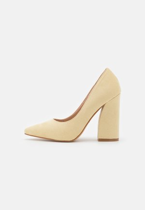 Zapato de tacón alto de ante beige con punta puntiaguda y tacón grueso, que presenta una textura suave y un diseño minimalista.