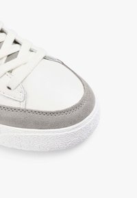 Zapatilla blanca con acentos de ante gris, con punta redonda, suela de goma texturizada blanca y cordones planos. Diseño limpio y minimalista.